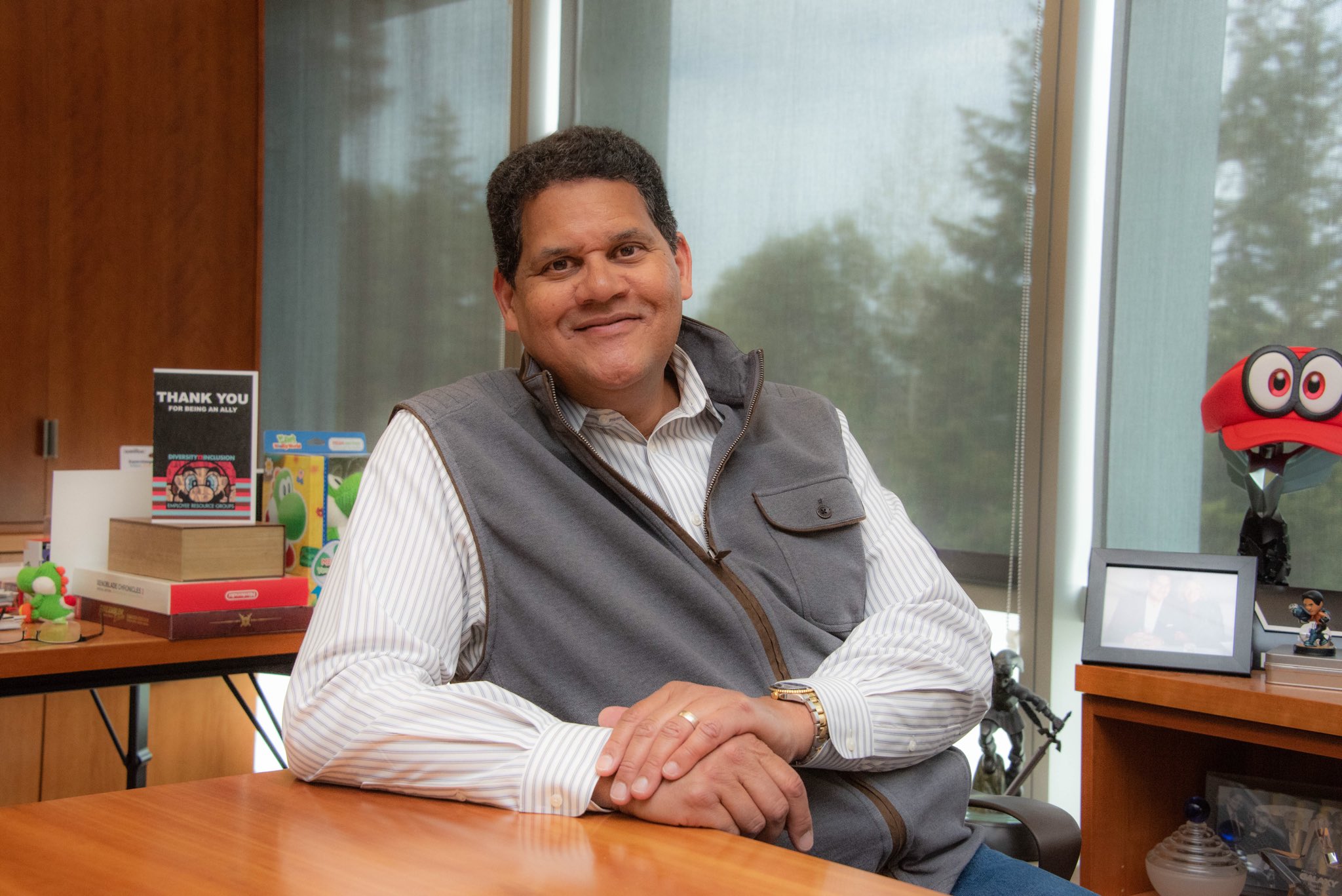 Reggie Fils-Aime sitting at his desk, smiling. There is Nintendo paraphernalia on the shelves behind him.