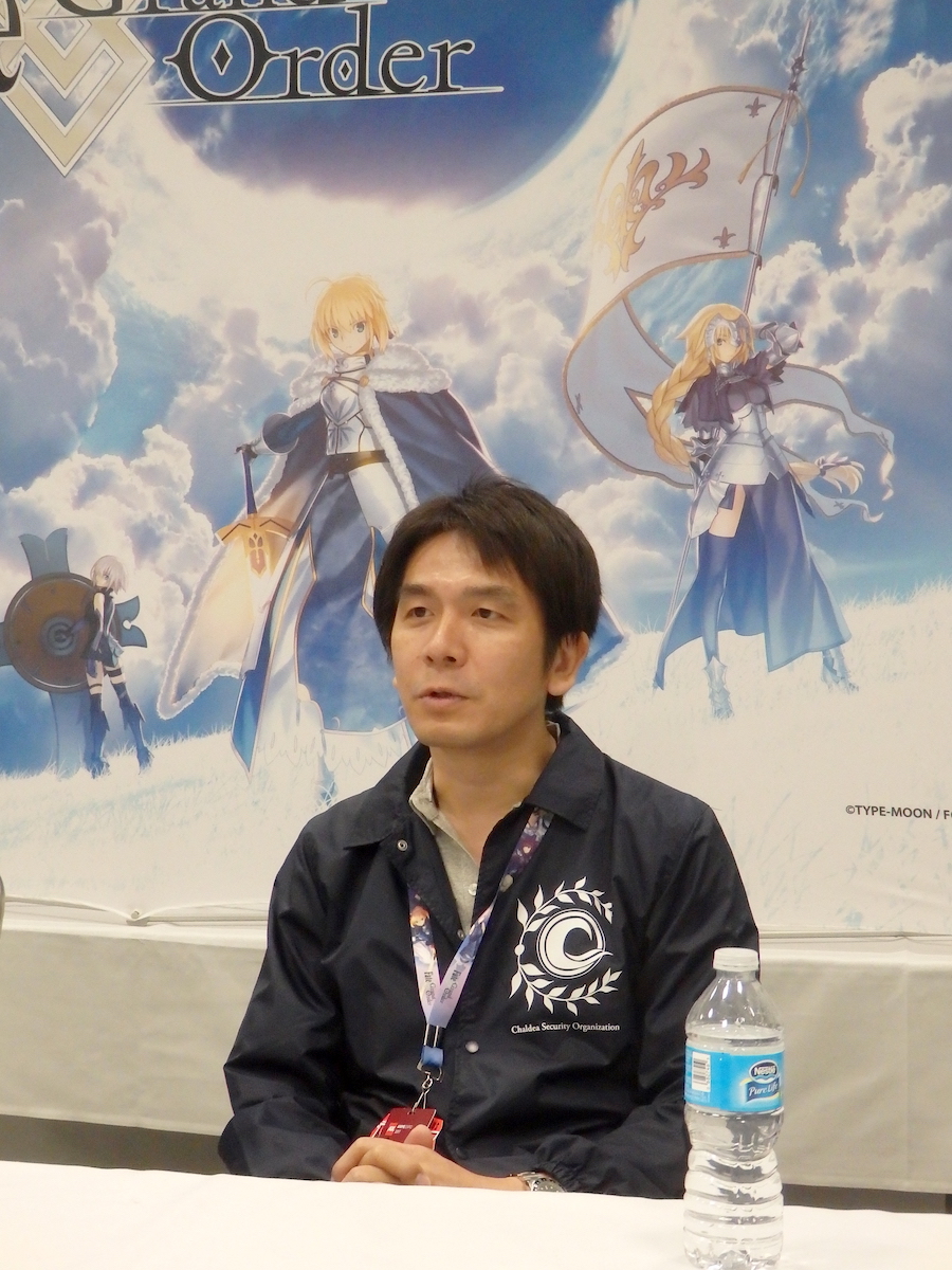 Yosuke Shiokawa speaking in front of a Fate/Grand Order banner.