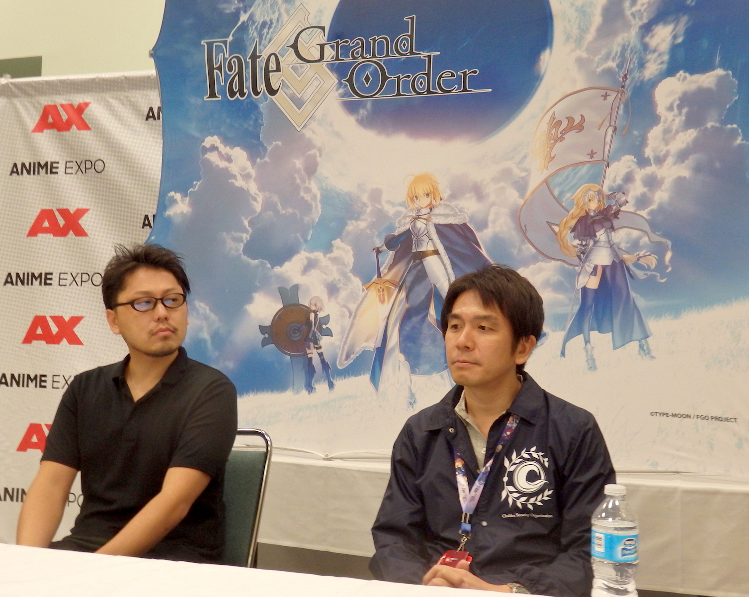 Two Japanese men sitting at a table in front of a banner for Fate: Grand Order.