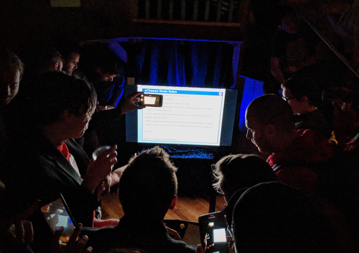 Game developers crowding around a screen playing E-Cheese Zone in a dark room at a party