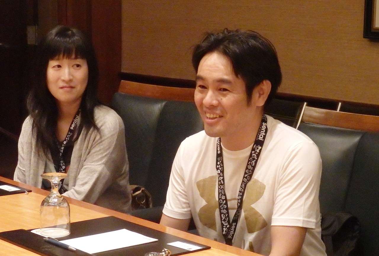 A Japanese woman and man sitting at a table. The main is smiling and talking.