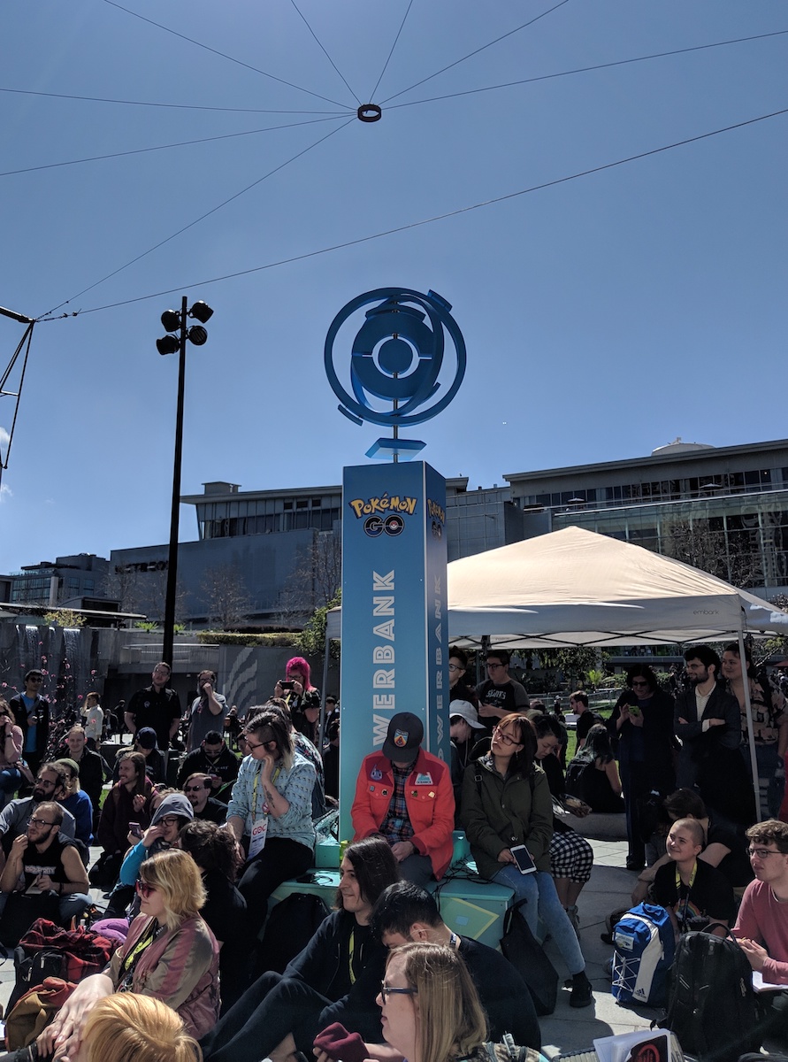 Crowd of young people sitting on the ground outside. In the center is a Pokémon Go-branded tower labeled 