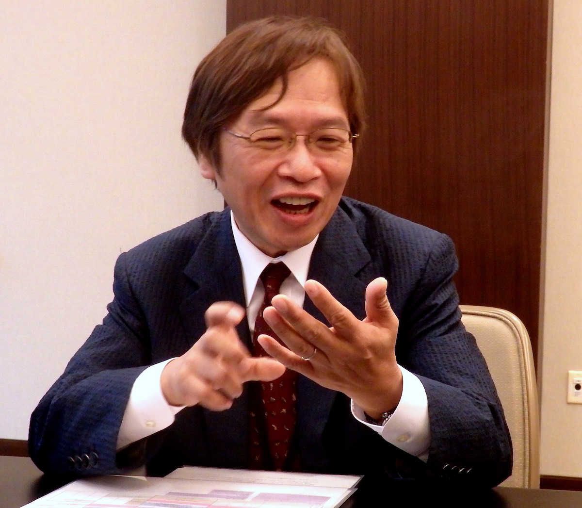 Katsuyuki Sumizawa sitting at a table, laughing and gesturing with both hands.
