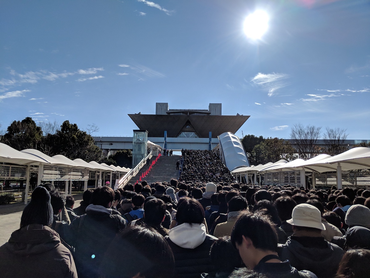 Massive crowd of hundreds of people walking down a pathway and up a stairwell toward the Tokyo Big Sight. There are blue skies overhead with a few wispy clouds.