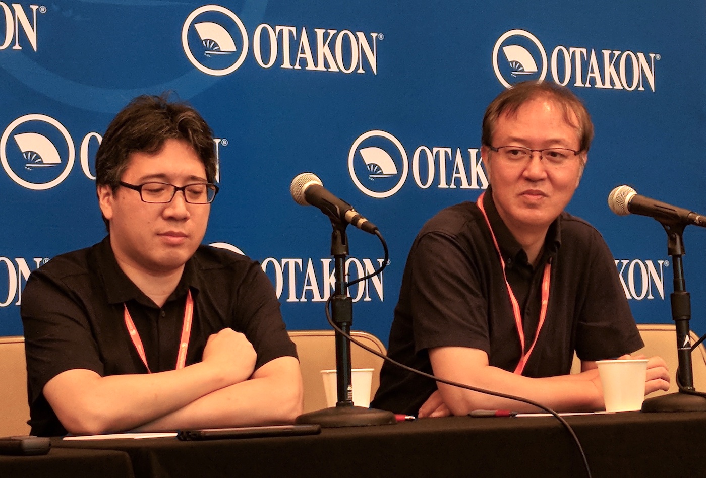 Yoshinari and Otsuka sitting in front of an Otakon banner, thinking