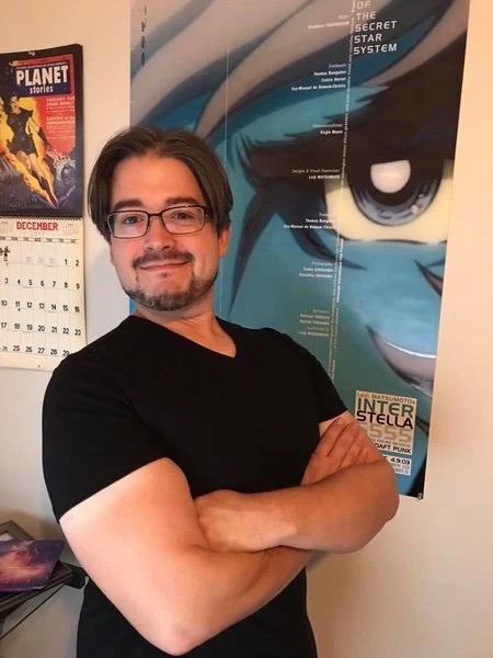 Zac Bertschy smiling with his arms folded in front of a poster from Interstella 5555.